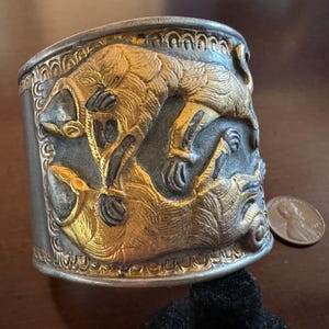 Könnte beinhalten: Silberarmband mit einem detaillierten Design aus zwei vergoldeten Tieren, möglicherweise Wölfen oder Hunden, die sich gegenüberstehen. Das Armband hat eine strukturierte Oberfläche und einen dekorativen Rand.