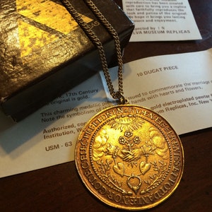 Puede incluir: Un collar con un colgante redondo de color dorado, adornado con un medallón detallado que representa dos manos entrelazadas con corazones y flores. El medallón está inscrito con el texto "PRBEEDNDN HYENFISSEKA SERENISSETTO ONCHAINGLO".