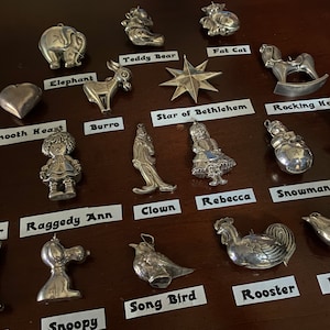 Könnte beinhalten: Eine Sammlung silberfarbener Weihnachtsschmuckstücke, darunter ein Elefant, ein Teddybär, ein Stern, ein Schaukelpferd und ein Schneemann. Weitere Ornamente sind ein Clown, ein Hahn und eine Friedenstaube. Jedes Ornament hat eine Schlaufe zum Aufhängen.