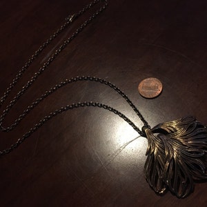 May include: A gold and black necklace with a large, ornate pendant shaped like a stylized feather. The pendant is made of metal and has a textured surface. The necklace is on a dark wooden surface.