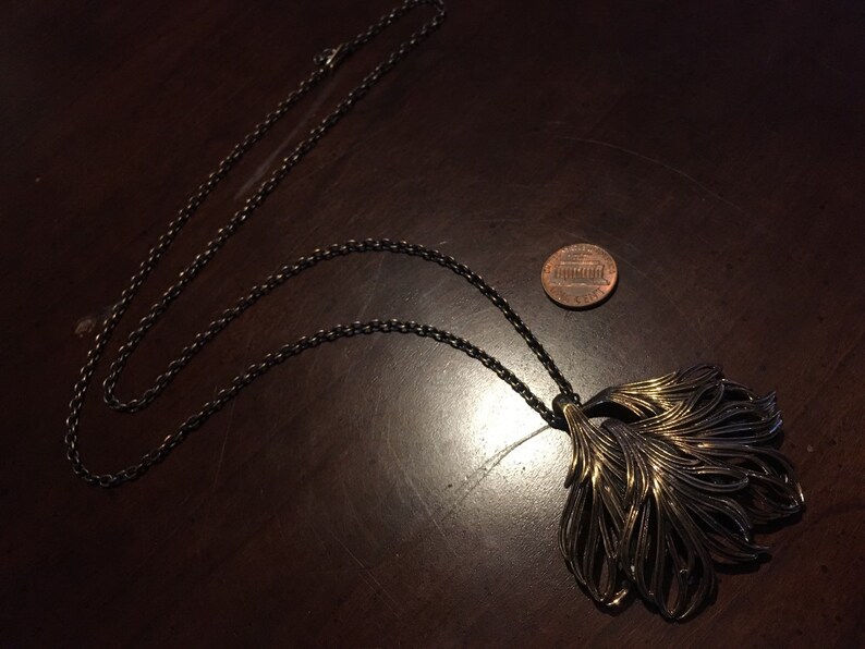 May include: A gold and black necklace with a large, ornate pendant shaped like a stylized feather. The pendant is made of metal and has a textured surface. The necklace is on a dark wooden surface.