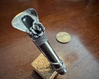 May include: A silver-toned metal hair accessory featuring a wide, flat, triangular top section and a cylindrical body. The piece is decorated with a sculpted rose motif and two bands of braided metal texture. A copper coin is shown for scale.