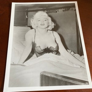 May include: A black and white photograph of a woman in a black lace bra lying in bed. She is looking directly at the camera with a serious expression. The bed is covered in white sheets and the background is a blurry image of a room.