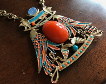 Puede incluir: Un collar de tono dorado con un gran colgante que presenta dos pájaros estilizados con esmalte naranja, azul y negro. El colgante también tiene una gran piedra naranja en cabujón en el centro.
