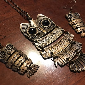 Könnte beinhalten: Ein goldfarbener Eulen-Halsketten- und Ohrring-Set. Die Halskette hat einen großen Eulen-Anhänger mit schwarzen Augen und filigranen Details. Die Ohrringe sind kleinere Eulen-Designs mit schwarzen Augen.