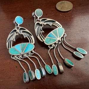 May include: Silver-tone earrings with turquoise details. Each earring has a wing-like design, an oval turquoise centrepiece, and dangling turquoise teardrops. The earrings are displayed on a wooden surface.