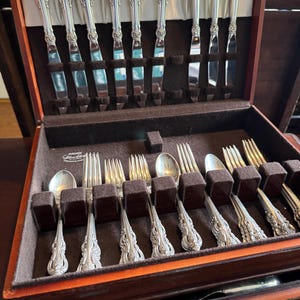 May include: A collection of silver cutlery, including forks, spoons, and knives, presented in a wooden case with a dark brown velvet interior. The silverware features intricate floral designs on the handles.