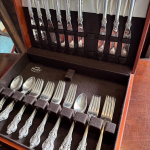 Può includere: Una scatola di legno per posate con un set di utensili d'argento. Il set include coltelli, forchette e cucchiai, tutti con dettagliati motivi floreali sui manici. La scatola è aperta, rivelando le posate.