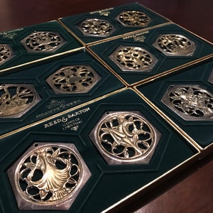 May include: Twelve silver and gold Christmas ornaments in their original boxes. Each ornament features a different design, including a swan, a tree, and a bell. The boxes are green with gold trim and feature the Reed & Barton logo.