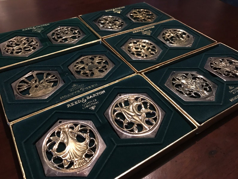 May include: Twelve silver and gold Christmas ornaments in their original boxes. Each ornament features a different design, including a swan, a tree, and a bell. The boxes are green with gold trim and feature the Reed & Barton logo.