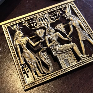 May include: A gold-toned metal brooch depicting an Egyptian scene with three figures. The central figure is seated on a throne, while the figures on either side stand. The scene is framed by a decorative border.