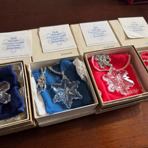 May include: A collection of six boxed Gorham full lead crystal snowflake ornaments. Each ornament is displayed in a velvet-lined box with a silver chain and a snowflake pendant. The boxes are white with gold lettering and the years 1975, 1976, 1977, 1978, 1979, and 1980 printed on the front.