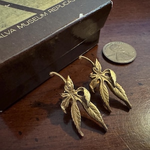 Puede incluir: Un par de pendientes dorados con forma de hoja sobre una superficie de madera oscura, junto a una caja con la inscripción "ALVA MUSEUM REPLICAS" y una moneda. Los pendientes miden unos 5 cm.