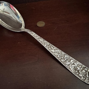 May include: A large silver serving spoon with a polished oval bowl and a long handle featuring an intricate embossed floral pattern. The handle displays detailed carvings of various flowers and leaves, and a small round coin is placed nearby.