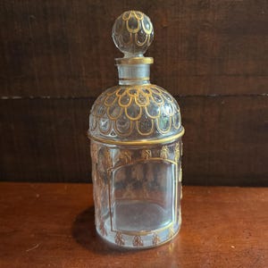 May include: A clear glass decanter featuring a rounded stopper and a cylindrical body. The glass is decorated with gold-coloured painted details, including a scalloped pattern on the shoulder and ornate, symmetrical motifs around the main body.