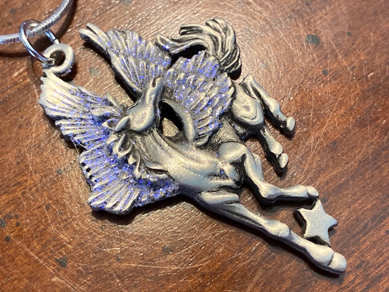 May include: A silver-toned Pegasus pendant with blue glitter accents on its wings, attached to a silver chain. The mythical creature is depicted mid-flight, with a star at the end of its leg. The pendant is a jewelry item.