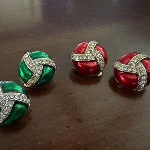May include: A set of four clip-on earrings. Two earrings are green with a white rhinestone design and two earrings are red with a white rhinestone design.