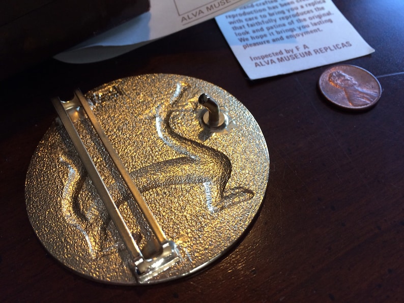 May include: A silver-toned metal belt buckle with a detailed design of a mythical creature. The buckle has a hinged pin and a loop for attaching to a belt.
