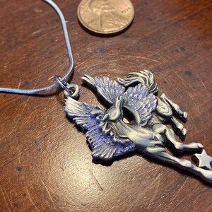 May include: A silver-toned Pegasus pendant necklace with blue accents. The pendant features a winged horse design with a star at the end. The necklace is on a silver chain, and a penny is next to the pendant.