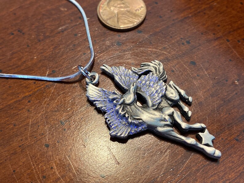 May include: A silver-toned Pegasus pendant necklace with blue accents. The pendant features a winged horse design with a star at the end. The necklace is on a silver chain, and a penny is next to the pendant.