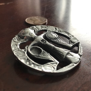 May include: A silver Celtic knot brooch featuring an intricate design of a woman holding a bird in each hand. The brooch is circular and has a textured finish.