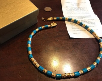 May include: A turquoise and gold necklace with a clasp in the shape of a bird's head. The necklace is made of beads and is on a wooden surface. The text on the paper reads: "The Metropolitan Museum of Art 09-02611-3"