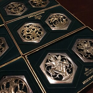 May include: Eight silver and gold Christmas ornaments in their original boxes. The ornaments are octagonal and feature intricate designs of fairies, flowers, and other motifs. The boxes are green with gold trim and feature the Reed & Barton logo and the text "125 Years of Christmas".