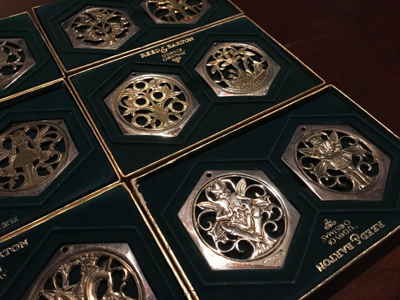 May include: Eight silver and gold Christmas ornaments in their original boxes. The ornaments are octagonal and feature intricate designs of fairies, flowers, and other motifs. The boxes are green with gold trim and feature the Reed & Barton logo and the text "125 Years of Christmas".