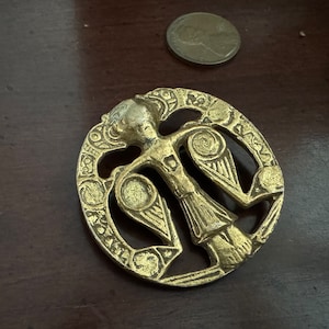 May include: A gold-coloured metal pendant featuring a stylised figure with wings, surrounded by a circular design with intricate details.