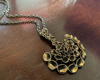 May include: A gold-toned necklace with a large, abstract pendant made of many small, rounded shapes. The pendant is attached to a silver chain.