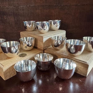 Könnte beinhalten: Eine Sammlung silberfarbener Metallschalen mit gehämmerten Texturen. Die Schalen sind auf Holzplattformen und einer dunkelbraunen Oberfläche angeordnet. Die Schalen haben eine reflektierende Oberfläche und sind unterschiedlich groß.