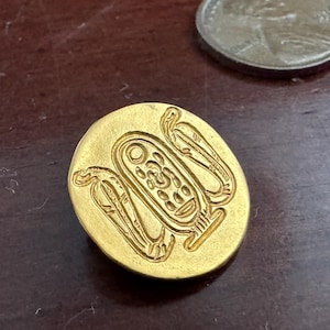 May include: A circular gold-toned button featuring an embossed Egyptian-style cartouche design flanked by two bird figures. The button is positioned next to a United States quarter coin for scale, showing the relative size of the metallic object.