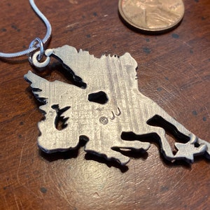 May include: Silver-toned pendant necklace featuring a detailed outline of a landmass, possibly a state or region. The pendant is attached to a thin, silver chain. The pendant has a textured surface and the initials "JJ" are visible.