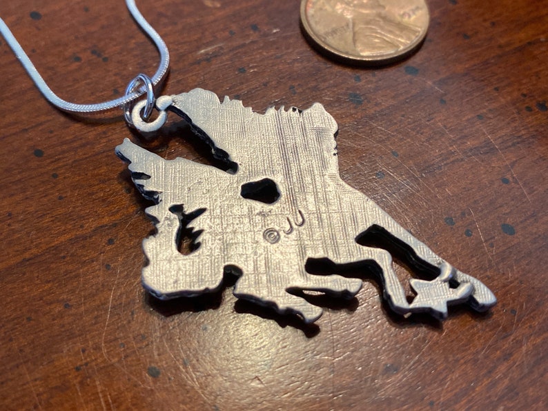 May include: Silver-toned pendant necklace featuring a detailed outline of a landmass, possibly a state or region. The pendant is attached to a thin, silver chain. The pendant has a textured surface and the initials "JJ" are visible.
