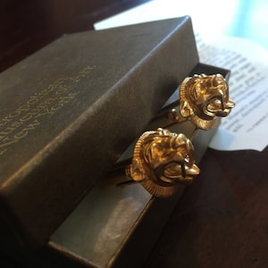 Pode incluir: Um par de botões de punho de ouro em forma de cabeças de leão, numa caixa castanha com o texto "The Metropolitan Museum of Art, New York" na tampa.