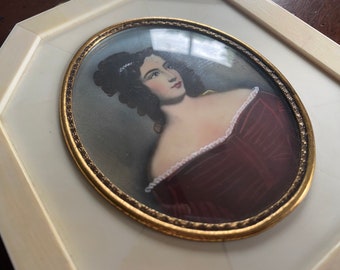 May include: A framed miniature portrait of a woman in a burgundy dress with a white lace trim. The portrait is set in a gold oval frame.
