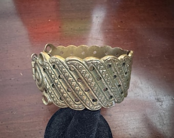 May include: A gold-toned metal bracelet with an intricate, woven design. The bracelet has a series of small, round holes throughout the design.