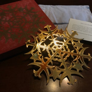 May include: A gold-coloured metal tree topper ornament in the shape of a snowflake with a star in the centre. The ornament is in a box with a white satin bag and a card that reads "Tree Top Ornament A galaxy of golden crescent moons and stars to grace your Christmas tree. It is a miniature copy of a piece by Ronald Bladen, a sculptor who explores complex relationships between form and space in his work.  The Metropolitan Museum of Art"