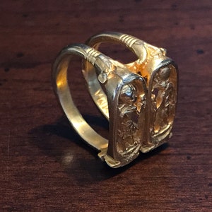 以下が含まれることがあります： 2 つのバンドが絡み合った金の指輪。それぞれにエジプト風のデザインが施され、人物とヒエログリフが描かれている。