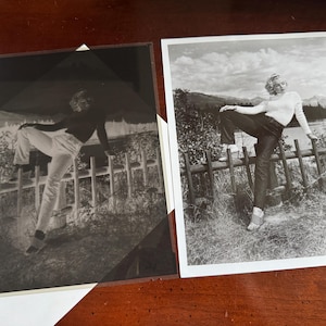 May include: Two black and white photographs of a person posing on a wooden fence. The person is wearing a white sweater, dark pants, and striped shoes. The background includes a landscape with trees and mountains. One photo is a negative.