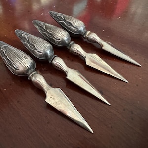 May include: Four silver-toned metal corn-shaped picks with pointed blades. Each pick features a textured, detailed corn cob handle with a smooth, tapered metal shaft and a sharp, triangular point, arranged in a diagonal row on a dark surface.