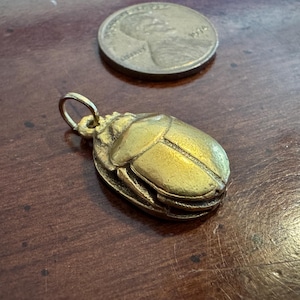 May include: A gold-toned metal beetle pendant featuring a detailed, segmented shell and a small circular bail at the top for attachment. The pendant is positioned near a United States penny for scale, showing the relative size of the insect charm.