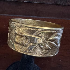 Puede incluir: Un brazalete ancho de color dorado con grabados en relieve detallados. El brazalete presenta un patrón repetitivo de follaje y figuras de animales. El metal tiene un aspecto ligeramente envejecido, lo que sugiere que es vintage o antiguo.