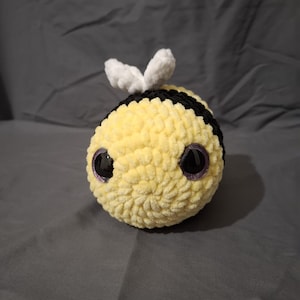 May include: A round, crocheted plush toy shaped like a bee, featuring a soft yellow body with horizontal black stripes. The bee has two small, white, textured wings on its back and two large, glossy, dark purple eyes positioned on the front.