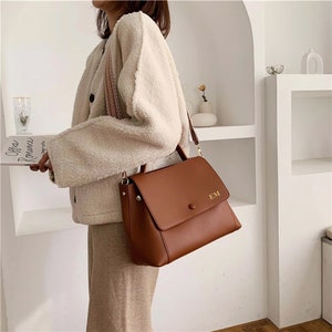 May include: A brown leather handbag with a flap closure and a shoulder strap. The bag has a top handle and gold-colored hardware. The initials "EM" are embossed on the front. The bag is being carried by a person wearing a cream-colored jacket.