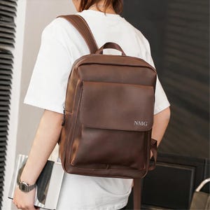 Może przedstawiać: Brązowy skórzany plecak z przednią kieszenią z klapką i regulowanymi paskami. Plecak ma wytłoczone inicjały "NMG" z przodu. Torba jest noszona na ramionach.