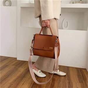 May include: A brown leather handbag with a top handle and a detachable, patterned shoulder strap. The bag features a flap closure with a button and gold-colored initials "EM". The strap has a cream and pink stripe design.