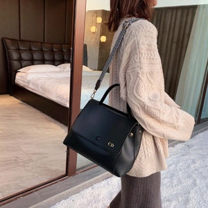 May include: A black leather satchel handbag with a gold buckle and a personalized monogram "CD". The bag has a long adjustable strap and a top handle.