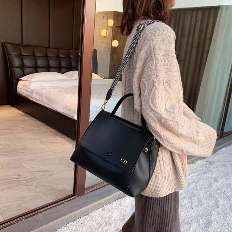 May include: A black leather satchel handbag with a gold buckle and a personalized monogram "CD". The bag has a long adjustable strap and a top handle.
