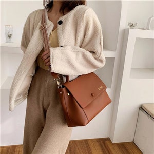 May include: A brown leather handbag with a flap closure and a shoulder strap. The bag has the initials "EM" on the front. The strap is a patterned beige and brown. The bag is being held by a person wearing a cream-colored jacket and tan pants.
